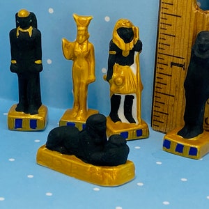 May include: Five small figurines of Egyptian deities, each standing on a gold base with blue accents. The figurines are painted in black, gold, and white.