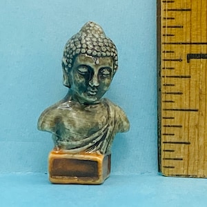 Puede incluir: Un pequeño busto de cerámica de Buda con un esmalte gris y verde. El busto está sentado sobre una base marrón.