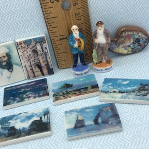 May include: A collection of miniature ceramic tiles featuring famous paintings by Claude Monet. The tiles depict scenes of water lilies, a cathedral, a tree, a bridge, and a seaside town. There are also two miniature figurines of Claude Monet, one holding a paint palette and the other holding a paintbrush.