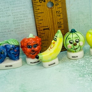 Peut inclure: Un ensemble de six figurines en céramique miniatures de fruits et légumes avec des visages. Les figurines sont peintes dans des couleurs vives et ont un style fantaisiste et cartoon. Les fruits et légumes comprennent une banane, un melon, une framboise, une prune, une prune et une pomme. Chaque figurine a une petite base avec le nom du fruit ou du légume écrit dessus en français.