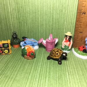 May include: A collection of miniature ceramic figurines depicting gardening themes. The figurines include a watering can, a wheelbarrow, a potted plant, a basket of vegetables, a turtle, a ladybug, a birdhouse, and a woman gardening.
