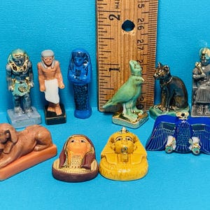 May include: A collection of ten miniature figurines depicting Egyptian gods and goddesses. The figurines are made of painted ceramic and feature intricate details. The figurines include a pharaoh, a sphinx, a hawk, a cat, and a goddess with a cobra.