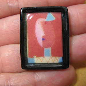 May include: A black framed resin cabochon with a colorful abstract image of a face with a blue hat and a red, white, and blue patterned background.