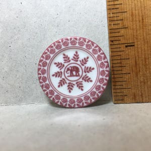 May include: A small, white ceramic plate with a pink floral border and a pink elephant design in the center. The elephant is surrounded by a pink floral design.