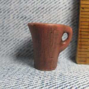May include: A small, red-brown ceramic jug with a handle. The jug has a textured surface and a slightly rounded shape.