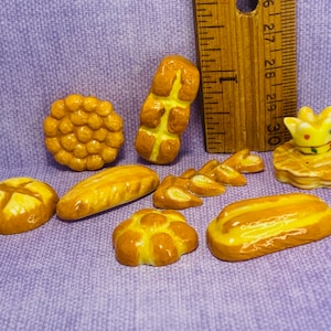 May include: A collection of miniature ceramic bread rolls, loaves, and pastries in various shapes and sizes. The pieces are painted in shades of yellow and brown, resembling freshly baked bread.