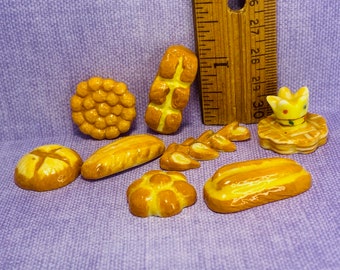 May include: A collection of miniature ceramic bread rolls, loaves, and pastries in various shapes and sizes. The pieces are painted in shades of yellow and brown, resembling freshly baked bread.