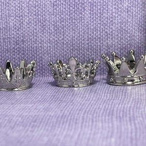 May include: Three miniature silver crowns with intricate details are arranged on a textured purple surface. The crowns vary in design, with one featuring a fleur-de-lis pattern. A wooden ruler is visible in the background, providing scale.