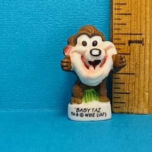 Può includere: Piccola statuina di Baby Taz, il personaggio dei Looney Tunes. La figura è marrone, bianca e rosa, con un ampio sorriso. La base è bianca con il testo "BABY TAZ TM & WBE (S07)".