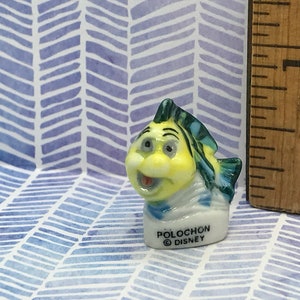 May include: A small ceramic figurine of a yellow and blue fish with a white base. The fish has a big smile and is wearing a blue and white scarf. The base of the figurine says "Polochon Disney".