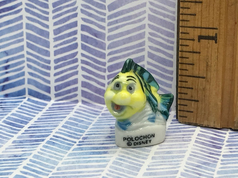 May include: A small ceramic figurine of a yellow and blue fish with a white base. The fish has a big smile and is wearing a blue and white scarf. The base of the figurine says "Polochon Disney".