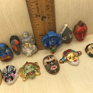 May include: Twelve miniature ceramic masks with various designs and colours, including blue, green, red, gold, and black. The masks are arranged on a wooden surface.