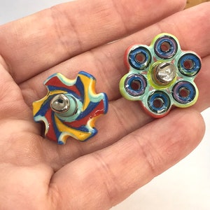 Puede incluir: Dos pomos de cerámica para cajones con acabado brillante. Uno presenta un patrón en espiral multicolor en rojo, amarillo, azul y blanco. El otro tiene forma de flor con seis pétalos circulares, cada uno con un anillo central de color azul.