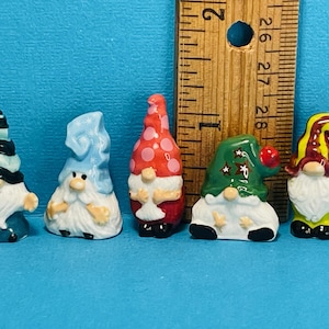 Peut inclure: Cinq nains de jardin miniatures en céramique avec des chapeaux et des barbes de différentes couleurs. Les nains sont debout sur une surface bleue à côté d'une règle.