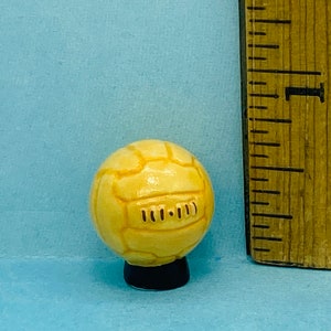 May include: A small, yellow ceramic soccer ball with black stitching and a black base. The ball is about 1 inch in diameter.