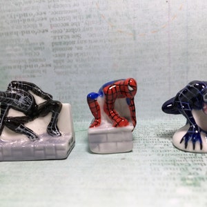 May include: Three small ceramic figurines of Spiderman in different poses. The figurines are painted in red, blue, and black. The figurines are on a white background.