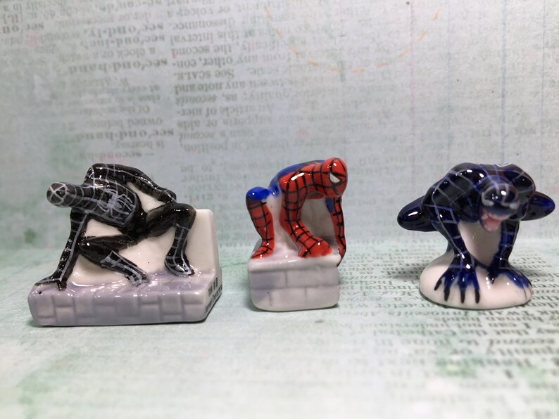 May include: Three small ceramic figurines of Spiderman in different poses. The figurines are painted in red, blue, and black. The figurines are on a white background.