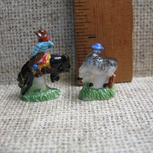 May include: Two miniature ceramic figurines. The first figurine is a cowboy riding a bucking horse. The cowboy is wearing a blue and white checked shirt, a brown hat, and brown trousers. The horse is black with a white mane and tail. The second figurine is a man wearing a blue hat and a grey shirt. He is standing on a green base.