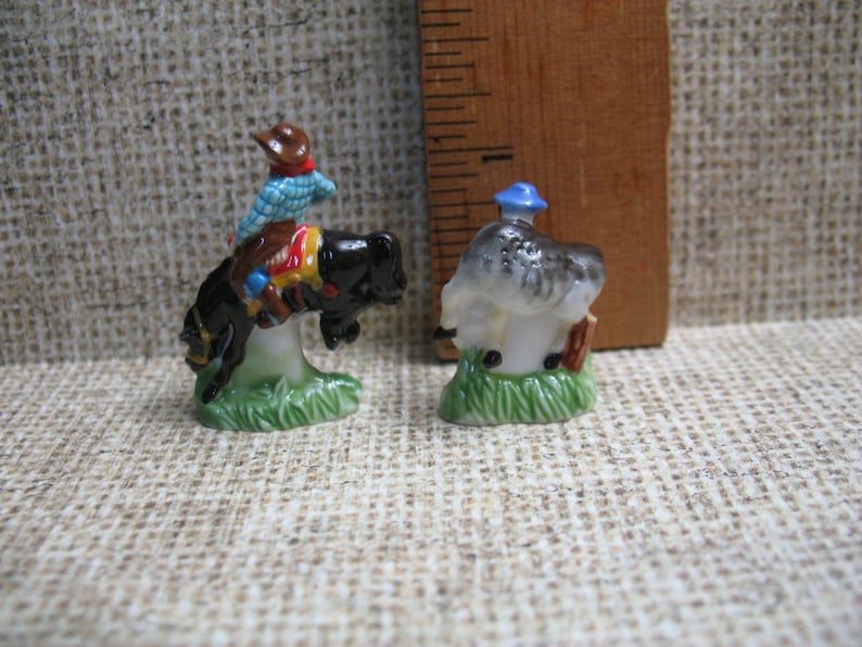 May include: Two miniature ceramic figurines. The first figurine is a cowboy riding a bucking horse. The cowboy is wearing a blue and white checked shirt, a brown hat, and brown trousers. The horse is black with a white mane and tail. The second figurine is a man wearing a blue hat and a grey shirt. He is standing on a green base.