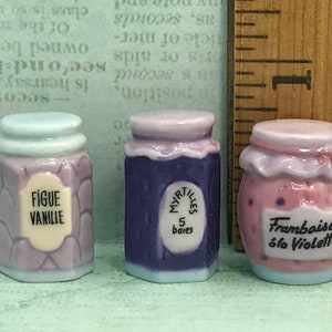 Op de afbeelding: Drie miniatuur keramische potten met deksels, elk versierd met een ander fruitontwerp. De potten zijn gelabeld met "Figue Vanille", "Myrtilles 5 baies" en "Framboise à la Violette".
