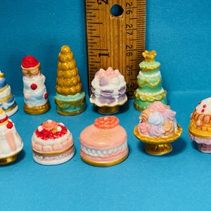 Puede incluir: Una colección de diez pasteles en miniatura de cerámica en varios colores y diseños. Los pasteles están decorados con glaseado, chispas y otros adornos. Están dispuestos sobre una superficie azul.