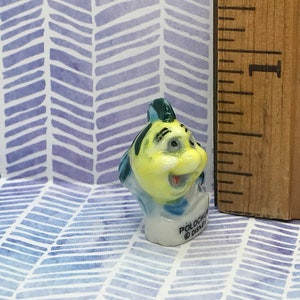 May include: A small, yellow and blue ceramic figurine of a fish with a smiling face. The figurine is about 1 inch tall and has the text "Polochon Disney" on the base.
