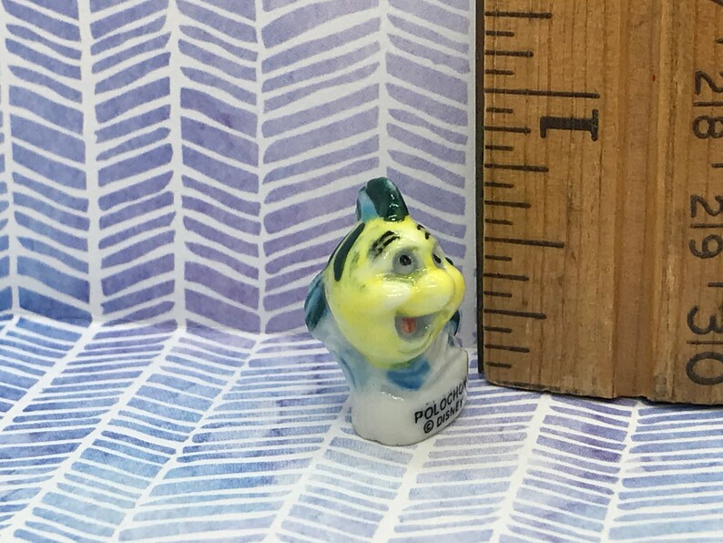 May include: A small, yellow and blue ceramic figurine of a fish with a smiling face. The figurine is about 1 inch tall and has the text "Polochon Disney" on the base.