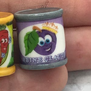 May include: A miniature cylindrical food can with a purple label featuring a cartoon plum character wearing a small gold crown and a green leaf. The label includes the text REINE CLAUDE in blue capital letters. The can has a grey top and bottom rim.