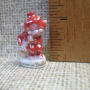 Puede incluir: Una pequeña figura de cerámica roja de un diablo sosteniendo un tenedor. La figura está de pie sobre una base blanca con el texto "comme un Diable" escrito en ella.