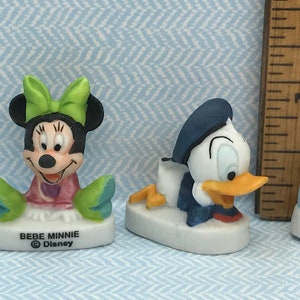 Może przedstawiać: Trzy małe ceramiczne figurki postaci z Disneya. Figurka Minnie Mouse w kolorze zielonym i r&oacute;żowym, figurka Donalda Kaczora w kolorze niebieskim i białym oraz figurka Myszki Miki w kolorze niebieskim. Każda figurka ma białą podstawę z napisem "BEBE" i symbolem praw autorskich Disneya.