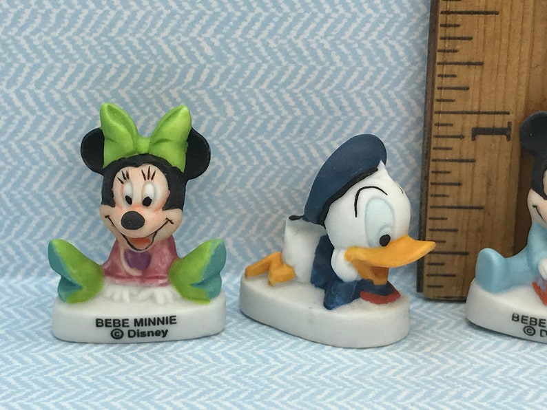Może przedstawiać: Trzy małe ceramiczne figurki postaci z Disneya. Figurka Minnie Mouse w kolorze zielonym i r&oacute;żowym, figurka Donalda Kaczora w kolorze niebieskim i białym oraz figurka Myszki Miki w kolorze niebieskim. Każda figurka ma białą podstawę z napisem "BEBE" i symbolem praw autorskich Disneya.