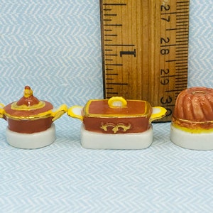 Op de afbeelding: Drie miniatuur porseleinen serveerschalen met deksels. De schalen zijn bruin met gouden accenten en gele rand. De schalen staan op witte voetjes.