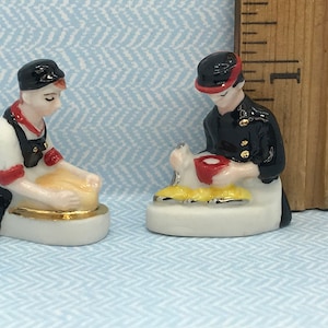 Puede incluir: Dos pequeñas figuritas de cerámica de personas. Una lleva un atuendo negro y rojo y sostiene un objeto redondo. La otra lleva un atuendo negro y rojo y sostiene un pequeño objeto blanco. Ambas figuritas están de pie sobre bases blancas.