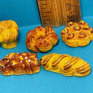 May include: Five miniature ceramic bread rolls in various shapes and colours. The rolls are glazed in shades of brown and yellow, with some featuring white or yellow accents.
