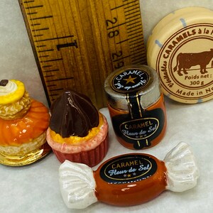 May include: A collection of miniature ceramic food items, including a chocolate and orange pastry, a yellow and orange pastry, a small jar of caramel with a label that reads "Caramel Fleur de Sel", and a brown and white candy with a label that reads "Caramel Fleur de Sel". There is also a small round container with a brown cow and the text "Caramels à la Fleur de Sel Véritables Poids net: 300 g Made in Normandie".