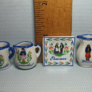 May include: Set of four miniature ceramic pieces with blue and white painted designs. The set includes a pitcher, a mug, and two smaller mugs. Each piece features a different design of a person in traditional clothing. The pitcher has the word "Yann" on it, one mug has the word "Erwan" on it, and the other mug has the word "Gwenadal" on it.