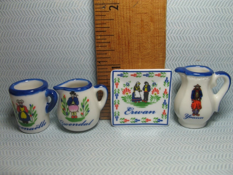 May include: Set of four miniature ceramic pieces with blue and white painted designs. The set includes a pitcher, a mug, and two smaller mugs. Each piece features a different design of a person in traditional clothing. The pitcher has the word "Yann" on it, one mug has the word "Erwan" on it, and the other mug has the word "Gwenadal" on it.