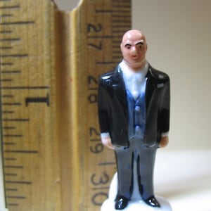 K&ouml;nnte beinhalten: Eine kleine, schwarz-blaue Figur von Lex Luthor aus DC Comics. Die Figur tr&auml;gt einen schwarzen Anzug mit einer blauen Weste und einem wei&szlig;en Hemd. Die Figur steht auf einem wei&szlig;en Sockel mit dem aufgedruckten Text "Lex Luthor TM & &copy; DC Comics (505)".