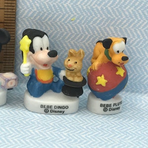 Może przedstawiać: Trzy ceramiczne figurki postaci z Disneya: Myszki Miki, Goofy'ego i Pluto. Myszka Miki ma na sobie niebiesko-biały str&oacute;j i trzyma niebiesko-białą zabawkę. Goofy ma na sobie niebiesko-ż&oacute;łty str&oacute;j i trzyma ż&oacute;łtą magiczną r&oacute;żdżkę w kształcie gwiazdy. Pluto ma na sobie ż&oacute;łto-pomarańczowy str&oacute;j i siedzi na czerwono-niebieskiej piłce z ż&oacute;łtymi gwiazdami.