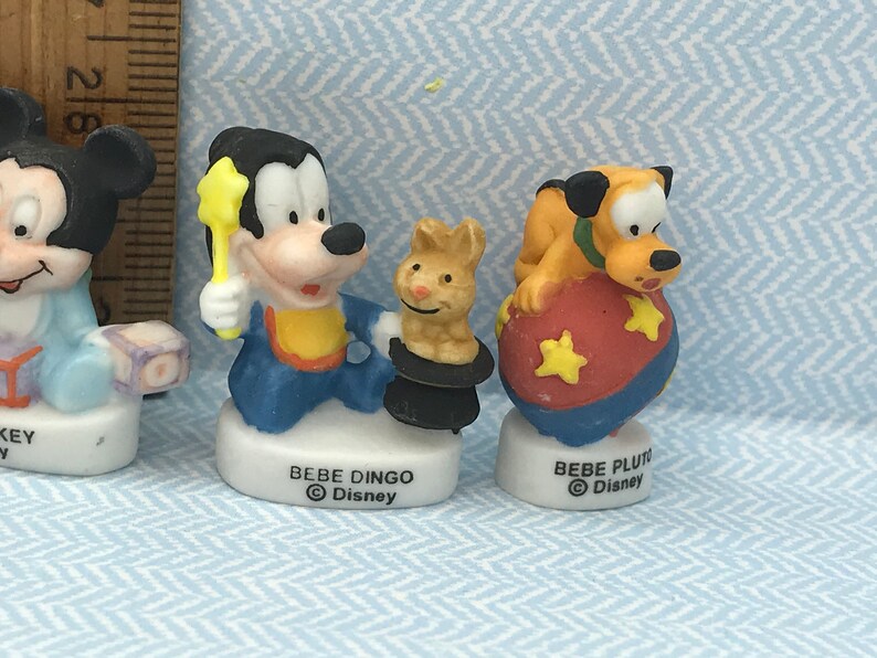 Może przedstawiać: Trzy ceramiczne figurki postaci z Disneya: Myszki Miki, Goofy'ego i Pluto. Myszka Miki ma na sobie niebiesko-biały str&oacute;j i trzyma niebiesko-białą zabawkę. Goofy ma na sobie niebiesko-ż&oacute;łty str&oacute;j i trzyma ż&oacute;łtą magiczną r&oacute;żdżkę w kształcie gwiazdy. Pluto ma na sobie ż&oacute;łto-pomarańczowy str&oacute;j i siedzi na czerwono-niebieskiej piłce z ż&oacute;łtymi gwiazdami.
