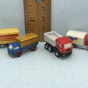 May include: Four miniature toy trucks made of ceramic. The trucks are in various colors, including yellow, brown, gray, red, and blue. The trucks have different styles of beds, including a flatbed, a dump bed, and a covered bed.