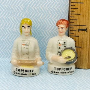 Puede incluir: Dos figuritas de cerámica de chefs del programa de televisión "Top Chef". Un chef sostiene un rodillo y el otro sostiene una sartén. Ambas figuritas llevan chaquetas de chef blancas con detalles negros y tienen el logotipo de "Top Chef" en sus chaquetas.