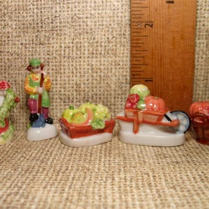 May include: Five miniature ceramic figurines depicting a farmer, a well, a wheelbarrow, a basket, and a cart, all filled with colorful vegetables. The figurines are painted in bright colors and have a whimsical, charming style.