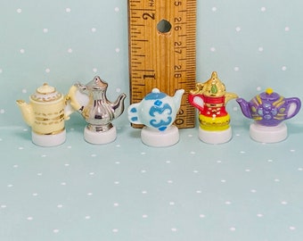 May include: Five miniature ceramic teapots in different colors and designs. The teapots are arranged in a row on a light blue background with white polka dots.