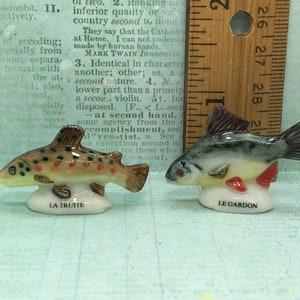 May include: Two miniature ceramic fish figurines, one labeled LA TRUITE and the other LE GARDON, positioned next to a wooden ruler showing a height of approximately 1.5 inches for the fish, featuring detailed painted scales and fins.