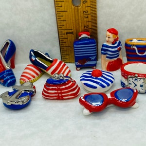 May include: A collection of miniature ceramic figurines in red, white, and blue with nautical and travel themes. The set includes a suitcase, a sailor girl, a hat, a pair of sunglasses, a purse, a pair of shoes, an anchor, a watch, and a beach bag.