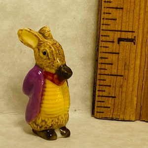 May include: A small ceramic figurine of a rabbit wearing a purple jacket, a yellow waistcoat, and a red neckerchief, positioned next to a wooden ruler showing the figure stands approximately 1.75 inches tall with a brown and white fur texture.