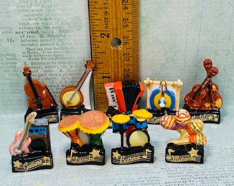 May include: A set of ten miniature musical instruments, each with a different design and color. The instruments include a violin, banjo, accordion, gong, guitar, drums, maracas, and a set of bongos. Each instrument is on a black base with a gold star and the name of the instrument written in gold letters.