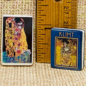 May include: Two small ceramic bookends featuring famous paintings by Gustav Klimt. The first bookend depicts "The Kiss" with two figures embracing in a mosaic-like pattern. The second bookend features "Judith and Holofernes" with a woman holding a sword and a severed head. Both bookends have a blue background with gold trim.