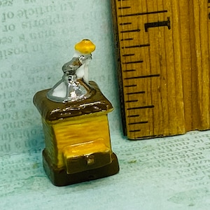 May include: A miniature ceramic coffee grinder with a yellow lid and brown base. The grinder has a silver handle and a white porcelain spout.